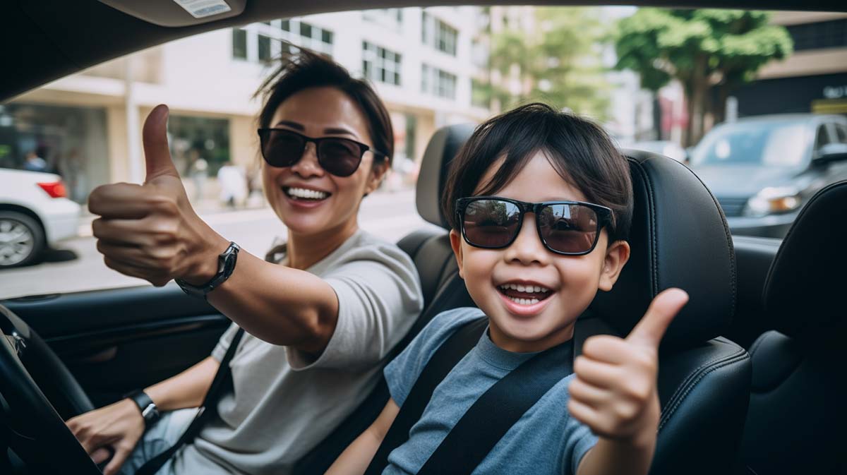 A woman smiling and driving, with her little son also smiling and giving the thumbs up.